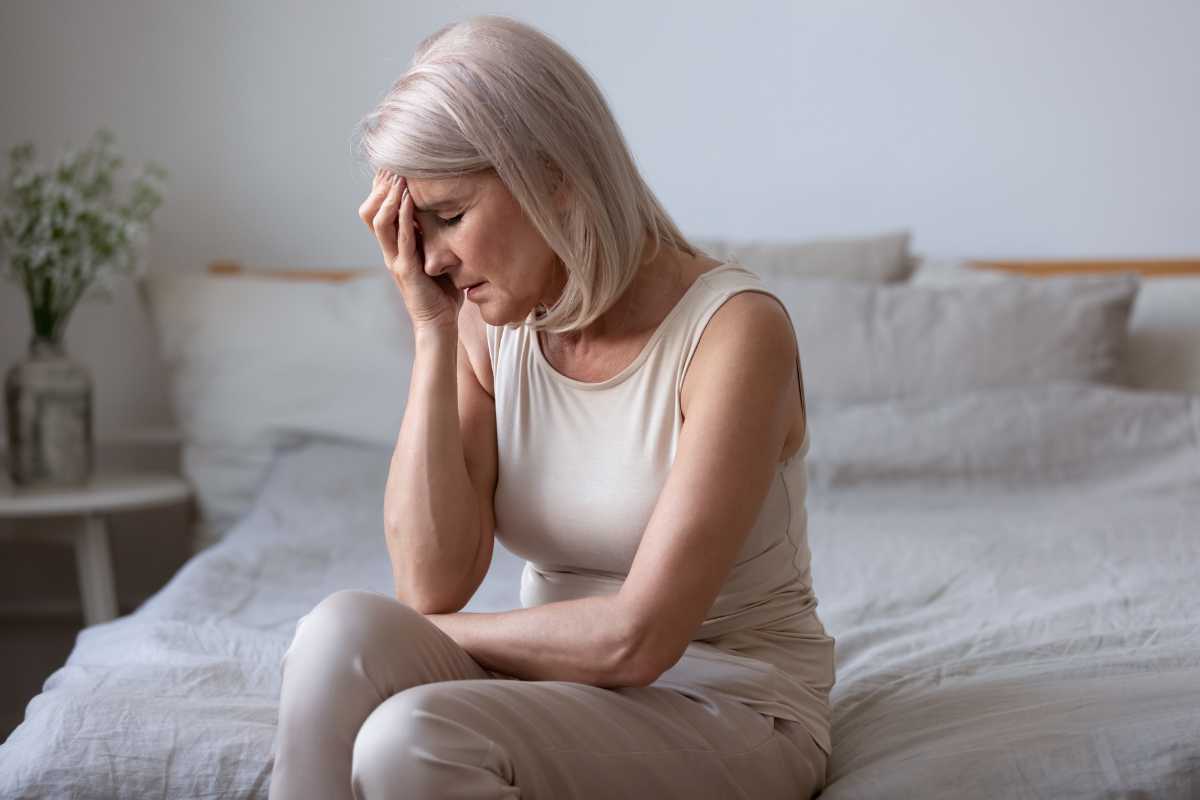 donna anziana seduta sul letto si tocca la testa con una mano